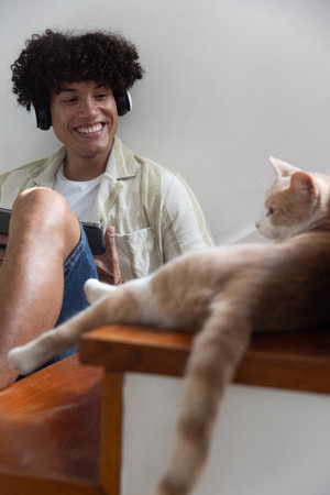 Man wearing headphones using tablet while sitting in living room next to cat on wooden table....