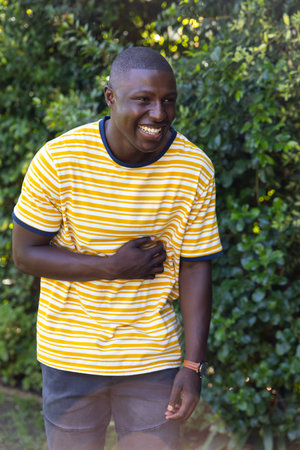 African American man laughing and glancing left in backyard wearing striped tee and wristwatch. Joy,...