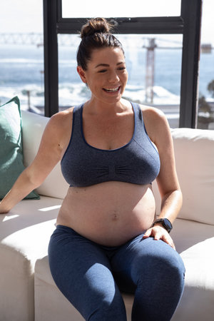 Pregnant woman sitting on sofa with green pillow wearing smartwatch by window overlooking ocean....