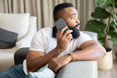 Mid adult African American man talking on smartphone while sitting on gray sofa in living room....