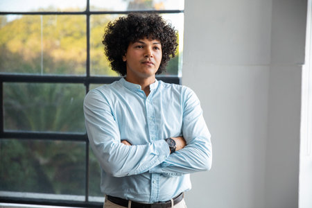 Man standing arms crossed wearing blue shirt, wristwatch, belt in studio by grid window....