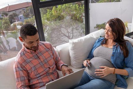 Diverse couple sitting on couch, man typing on laptop and pregnant woman holding ultrasound prints....