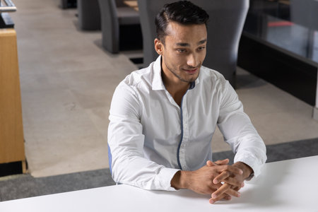 South Asian man sitting at white table with gray lounge chairs in office reception, copy space....