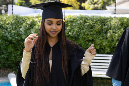 Female student wearing black gown adjusting mortarboard cap and standing on campus by white bench....