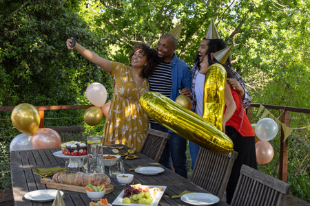 Diverse friends wearing party hats taking selfie on deck holding smartphone by table near balloons....