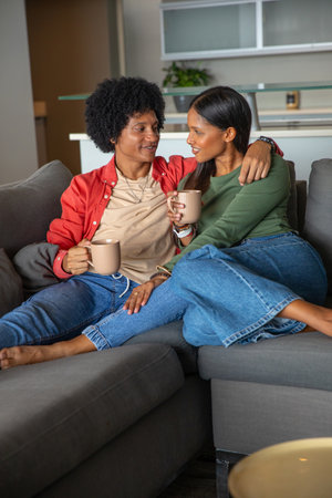 Diverse couple sitting on gray sofa in living room holding ceramic mugs with kitchen plant. Intimacy, relaxation, modern, cozy, lifestyle, home staging, warmthの写真素材