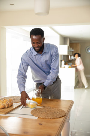 African American man placing loaf on woven placemat on table while partner using coffee machine. Home, breakfast, lifestyle, modern, domestic, brunch, morningの写真素材