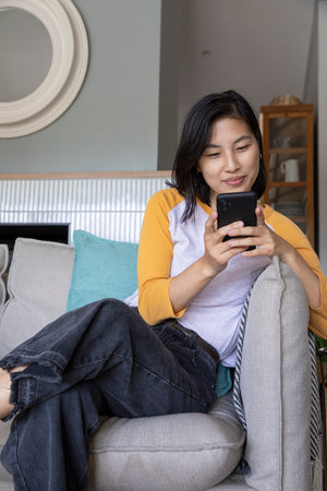 Asian woman sitting on gray couch at home holding black smartphone with wooden cabinet and mirror....