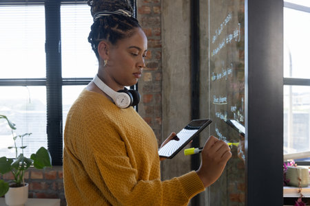 African American woman writing diagrams on glass board in studio with tablet and marker, copy space. Creative, innovative, workspace, contemporary, startup, collaboration, minimalisticの写真素材