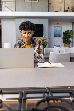 Asian man typing on laptop at kitchen island with stovetop burners, notebook and pen. Contemporary,...