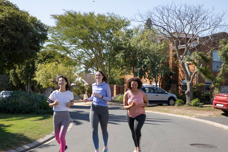 Diverse female friends jogging along suburban street wearing workout clothes and running shoes....