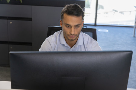 Hispanic male working at white desk in office using large monitor with mesh chair, storage cabinet....
