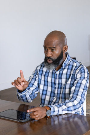 Mature African American man sitting at wooden table in home office wearing watch and using tablet. Professional, workplace, modern, concentration, wisdom, casual, communicationの写真素材