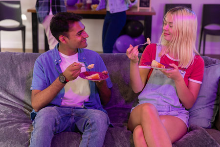 Diverse couple holding plates and forks eating cake on gray couch with purple balloons. Celebration,...