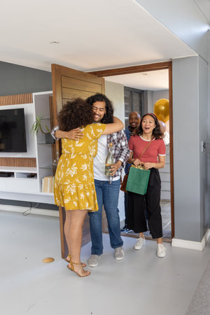 Diverse friends hugging couple at home entrance, holding wine bottle, gold balloons and gift bag....