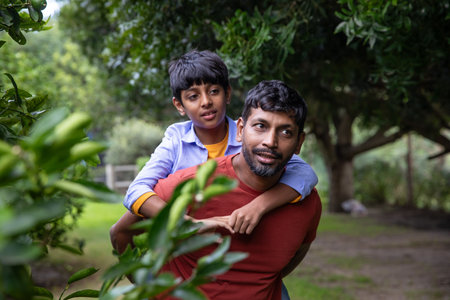 South Asian father and son giving piggyback ride while walking through lush orchard in soft light. Family, outdoors, nature, bonding, greenery, leisure, childhoodの写真素材