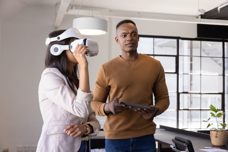 Diverse coworkers adjusting white VR headset and using tablet under hanging light fixture at office....