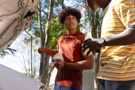 Diverse male friends cleaning dipstick with rag beside white car on suburban driveway under trees. Automotive, teamwork, casual, vintage, outdoor, friendship, maintenanceの写真素材