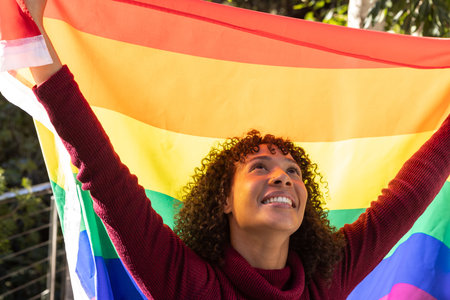 African American woman standing on balcony holding rainbow pride flag above head in sunlight....