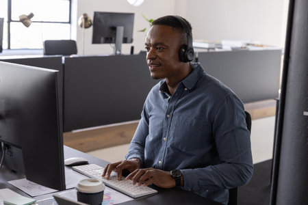 African American man typing on white keyboard at office desk with computer monitor and coffee cup....