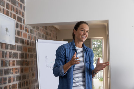 Woman gesturing while presenting graph on flip chart in lit meeting room wearing denim jacket....