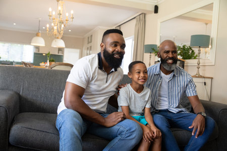 African American three-generation family sitting on gray sofa with chandelier near kitchen island....