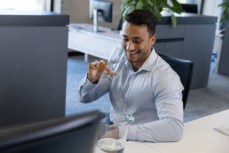 South Asian man sipping water from glass at office desk with computer monitor, copy space. Corporate, hydration, productivity, modern, professional, workspace, minimalistの写真素材