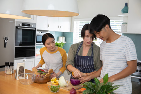 Asian family cooking together at kitchen island chopping vegetables and mixing salad in wooden bowl....