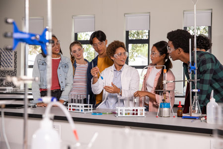 Diverse instructor wearing lab coat holding test tube while students leaning over lab bench....