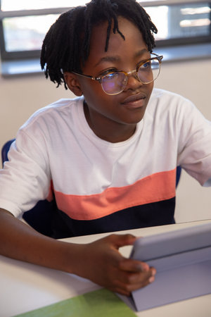 African American teen student sitting at desk in classroom wearing eyeglasses and holding tablet....