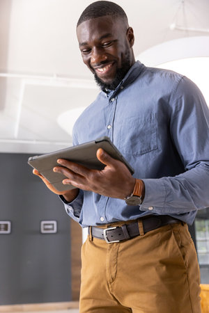 African American man standing in modern office holding tablet and wearing wristwatch, leather belt. Technology, professionalism, workspace, innovation, contemporary, corporate, communicationの写真素材