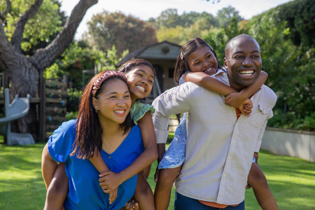 Diverse family giving piggyback rides on lush lawn in suburban backyard near wooden playset. Family, outdoor, recreational, happiness, leisure, casual, environmentの写真素材