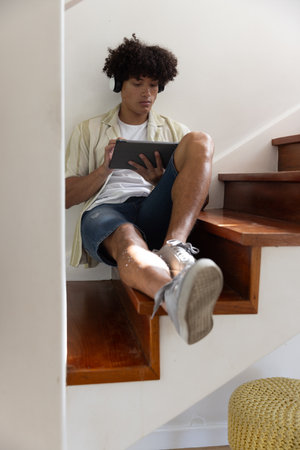 Male sitting on wooden stairs at home using tablet and wearing headphones near knitted pouf. Casual, relaxation, modern, comfort, technology, home, lifestyleの写真素材