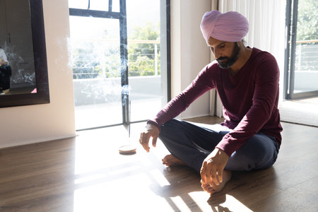 Indian man wearing turban and meditating at home by glass doors near incense holder, copy space....