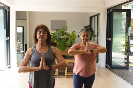 Female friends practicing yoga pose in home dining area with table, plant, fitness watches. Serenity, mindfulness, wellness, natural light, tranquility, healthy lifestyle, minimalistの写真素材
