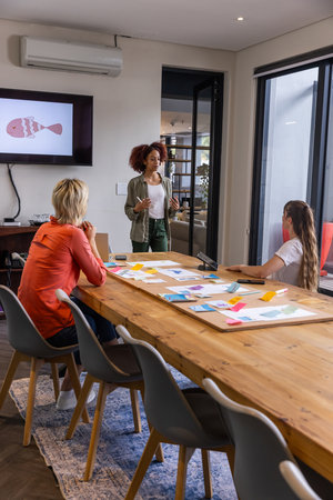 Diverse female coworkers collaborating around table with sticky notes and mockups in meeting room....