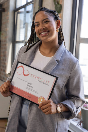 African American woman wearing blazer holding framed certificate in office by windows with plant....