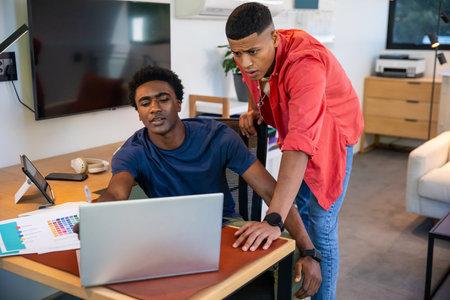 African American and Hispanic male colleagues collaborating on laptop with swatch in home office. Collaboration, teamwork, workspace, modern, productivity, innovation, technologyの写真素材