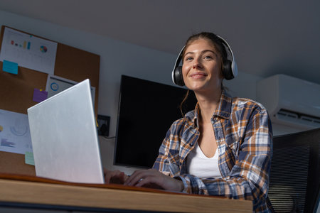 Woman wearing plaid shirt and headset typing on laptop under corkboard, monitor at home office desk....
