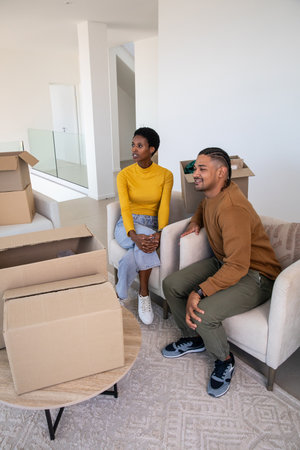 Diverse couple sitting on cream armchairs unpacking boxes on coffee table in modern living room....