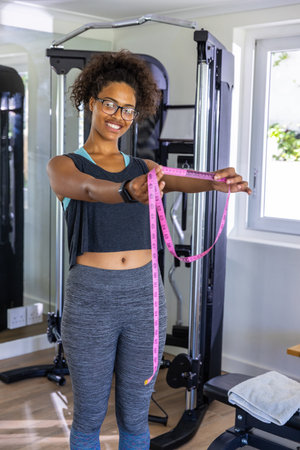 African American woman aged 29 in sportswear standing in home gym holding pink measuring tape....