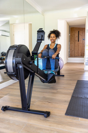 African American woman rowing on rowing machine in home gym with full-length mirror reflection....