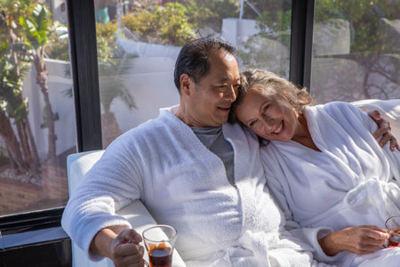 Diverse senior couple sitting on chair in sunroom wearing white bathrobes holding glass mugs of tea. Relaxation, intimacy, harmony, wellness, leisure, comfort, eleganceの写真素材