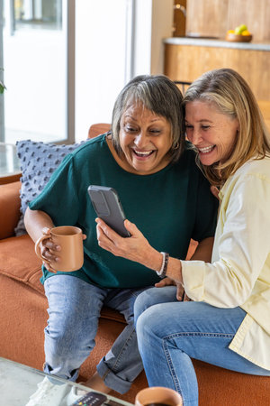 Diverse senior friends smiling and leaning toward smartphone holding mugs on sofa in living room. Companionship, leisure, cozy, relaxation, home, warmth, joyの写真素材