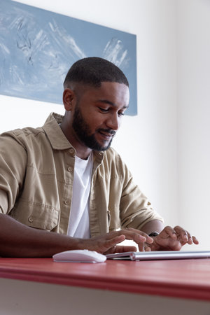 African American man wearing overshirt typing on tablet at red desk in office with wireless mouse....