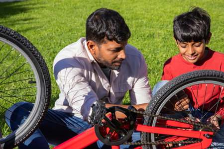 Indian father and son assembling red bicycle frame with screwdriver on sunny backyard lawn. Family, teamwork, craftsmanship, outdoor, mentorship, trust, recreationalの写真素材