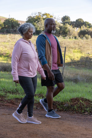 Senior African American couple walking dirt trail beside chain link fence wearing zip-up hoodies. Adventure, leisure, outdoor, exploration, companionship, rural, activeの写真素材