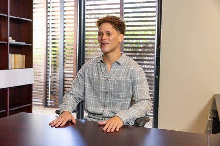 Man sitting at dark wood desk in well-lit office with wooden slats filtering light. Professional, workspace, minimalist, corporate, interior, productivity, modernの写真素材