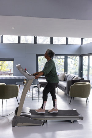 Woman walking on treadmill in modern living room near large windows framing green yard, copy space....