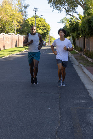 Diverse male friends running on suburban road wearing running shoes checking pace on sports watches....
